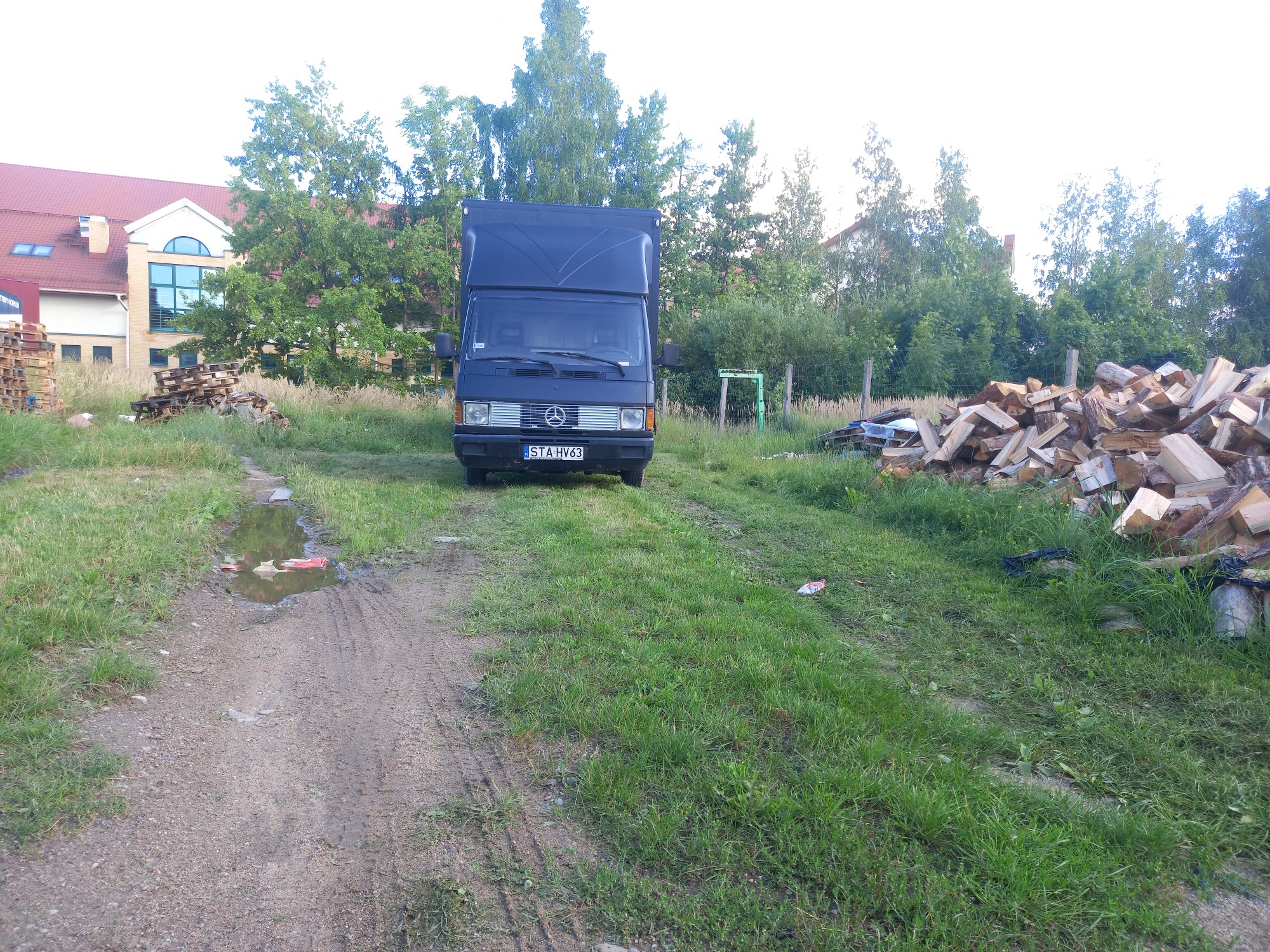 Czarny samochód dostawczy Mercedes zaparkowany na trawiastym terenie z pryzmami drewna w tle, widziany z przodu.