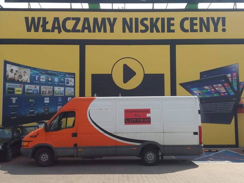 Pomarańczowo-biały samochód dostawczy z napisem 'Przeprowadzki P.J.' i numerem telefonu, zaparkowany na tle reklamy telewizorów i laptopów z hasłem 'Włączamy niskie ceny!'.