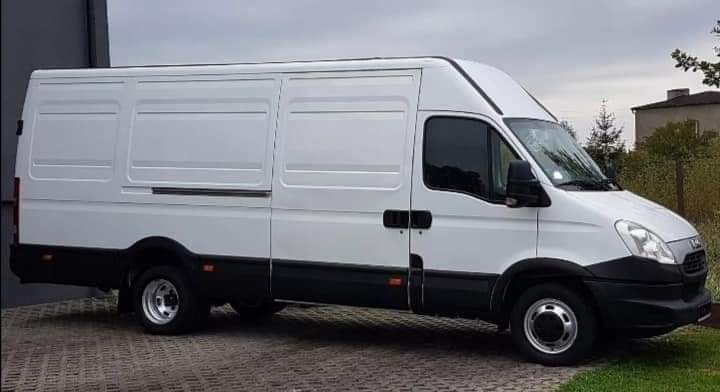 Biały samochód dostawczy typu furgon marki Iveco, zaparkowany na brukowanej powierzchni, z widocznym budynkiem w tle.