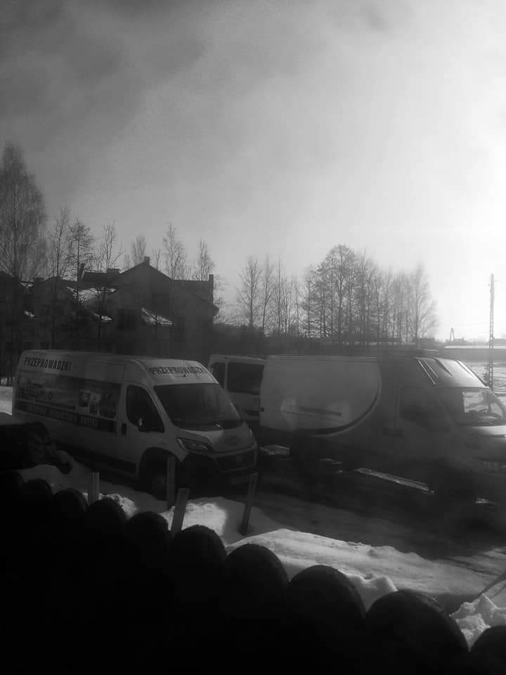 Czarne i białe zdjęcie kilku samochodów dostawczych zaparkowanych na śniegu, z logo firmy widocznym na jednym z nich, z budynkami i drzewami w tle pod pochmurnym niebem.