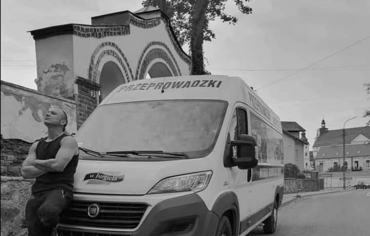 Mężczyzna oparty o białą furgonetkę z napisem 'Przeprowadzki' na dachu, w tle budynki miejskie i pochmurne niebo. Zdjęcie czarno-białe.