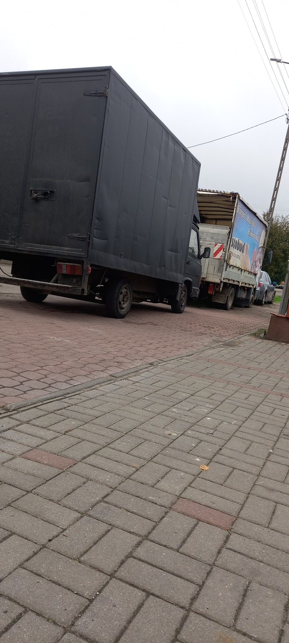 Dwa samochody dostawcze zaparkowane na brukowanej ulicy, jeden czarny i jeden z reklamą na plandece. Widoczny słup elektryczny i fragment drzewa w tle.