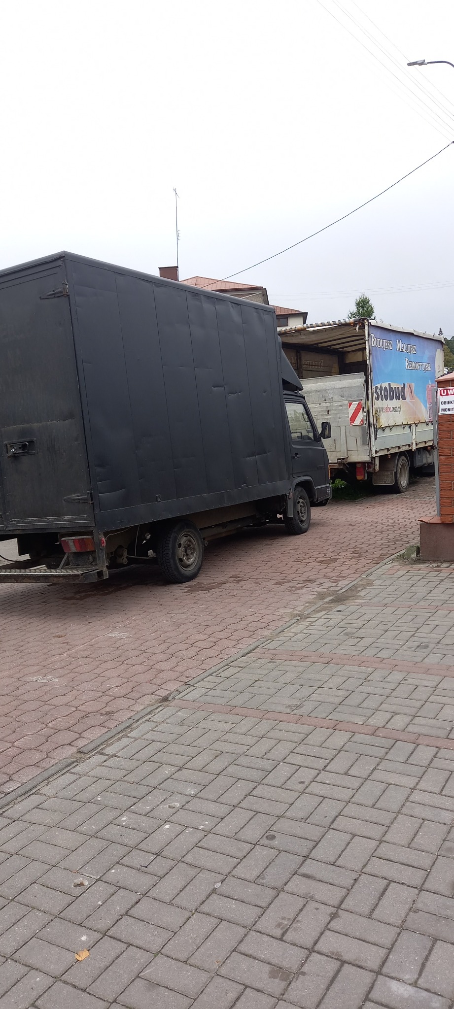 Dwa samochody dostawcze zaparkowane na brukowanej ulicy, jeden czarny z zamkniętą skrzynią ładunkową, drugi biały z reklamą na plandece.