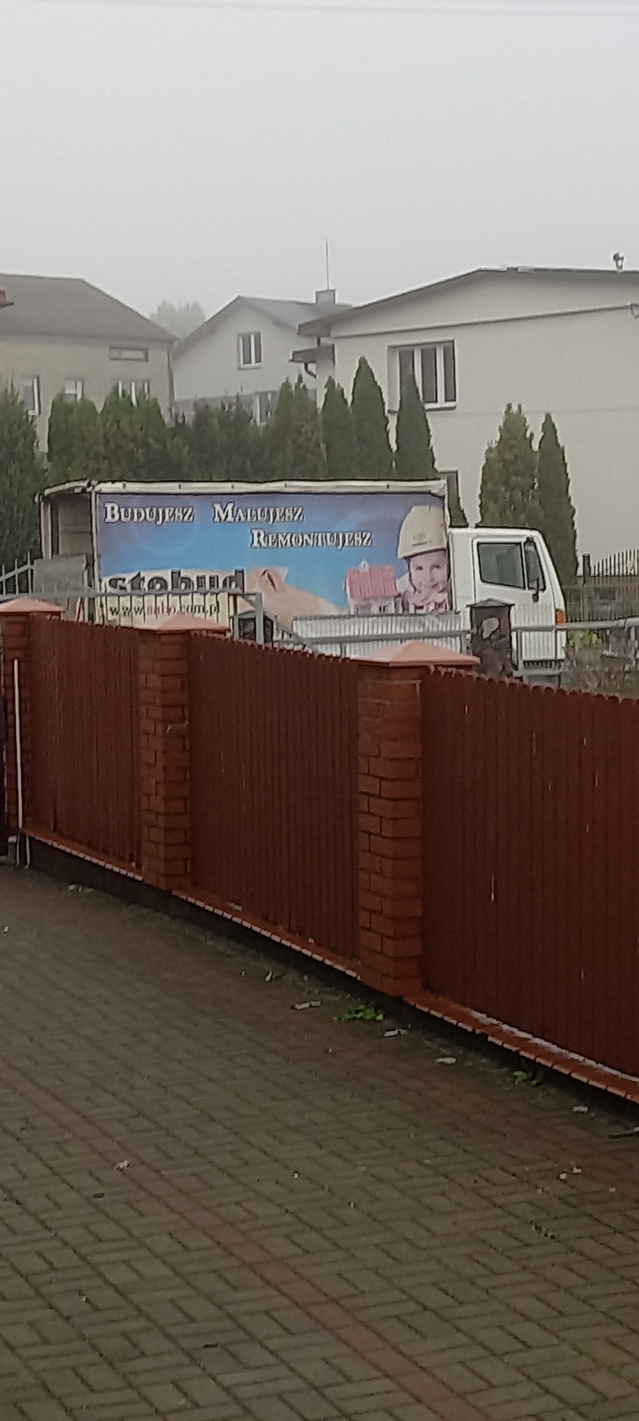 Ciężarówka z reklamą firmy budowlanej 'Stobud' z hasłem 'Budujesz, Malujesz, Remontujesz', zaparkowana na tle zabudowań mieszkalnych w pochmurny dzień.