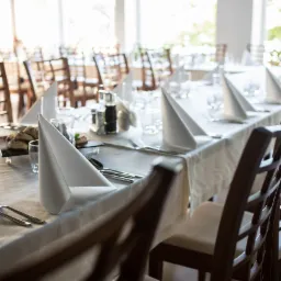 Elegancko nakryty st&oacute;ł z białym obrusem i serwetkami złożonymi w stożki, ciemne drewniane krzesła, szkło, sztućce i przekąski na czarnych talerzykach w jasnym wnętrzu restauracji.