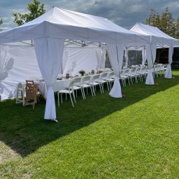 P.G.Połaniecki - Dwa białe namioty plenerowe z białymi zasłonami, ustawione na trawie, z długim stołem i krzesłami przygotowanymi do przyjęcia, widoczny drewniany fotelik dla dziecka.