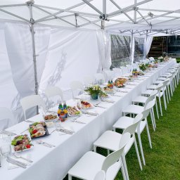 P.G.Połaniecki - Długi stół nakryty białym obrusem, ustawiony pod białym namiotem plenerowym, z rzędami białych krzeseł po bokach, zastawiony przekąskami, napojami i kwiatami.