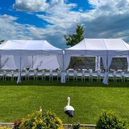 P.G.Połaniecki - Dwa białe namioty połączone ze sobą, ustawione na zielonym trawniku, przygotowane do przyjęcia z nakrytymi stołami i białymi krzesłami. Na pierwszym planie dekoracja ogrodowa w postaci bociana.