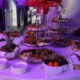 Stół z białym obrusem, na którym znajdują się patery z ciastami, owocami (winogrona, mandarynki) oraz ananas w dekoracyjnym koszyku. W tle widoczne srebrne beczki i drzwi.