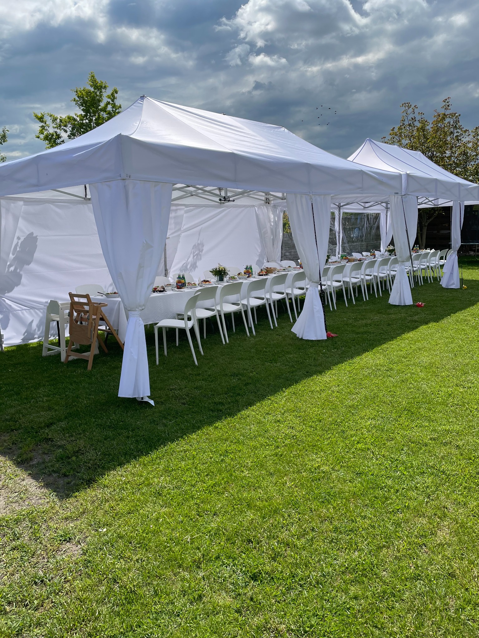 Dwa białe namioty plenerowe z białymi zasłonami, ustawione na trawie, z długim stołem i krzesłami przygotowanymi do przyjęcia, widoczny drewniany fotelik dla dziecka.
