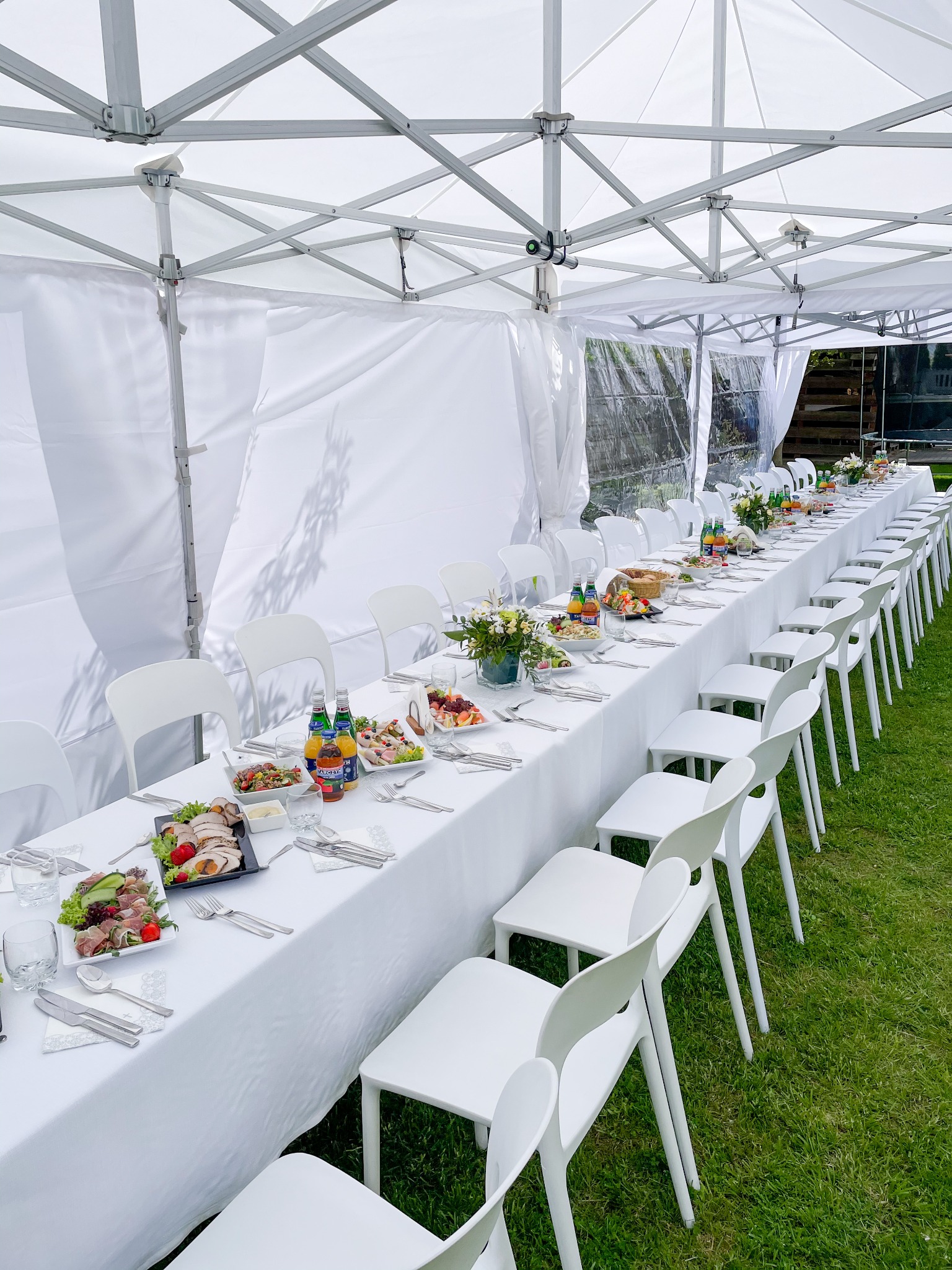 Długi stół nakryty białym obrusem, ustawiony pod białym namiotem plenerowym, z rzędami białych krzeseł po bokach, zastawiony przekąskami, napojami i kwiatami.
