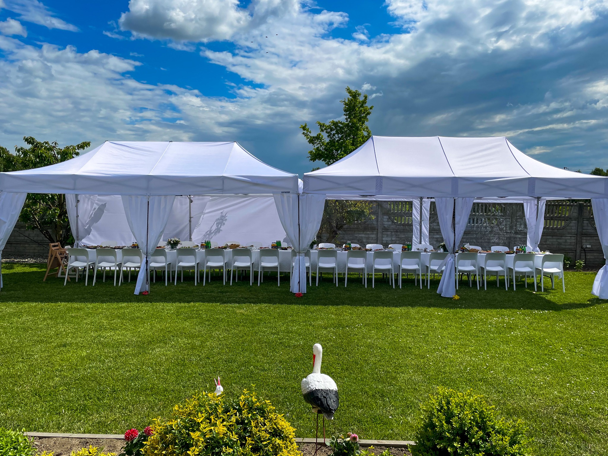 Dwa białe namioty połączone ze sobą, ustawione na zielonym trawniku, przygotowane do przyjęcia z nakrytymi stołami i białymi krzesłami. Na pierwszym planie dekoracja ogrodowa w postaci bociana.