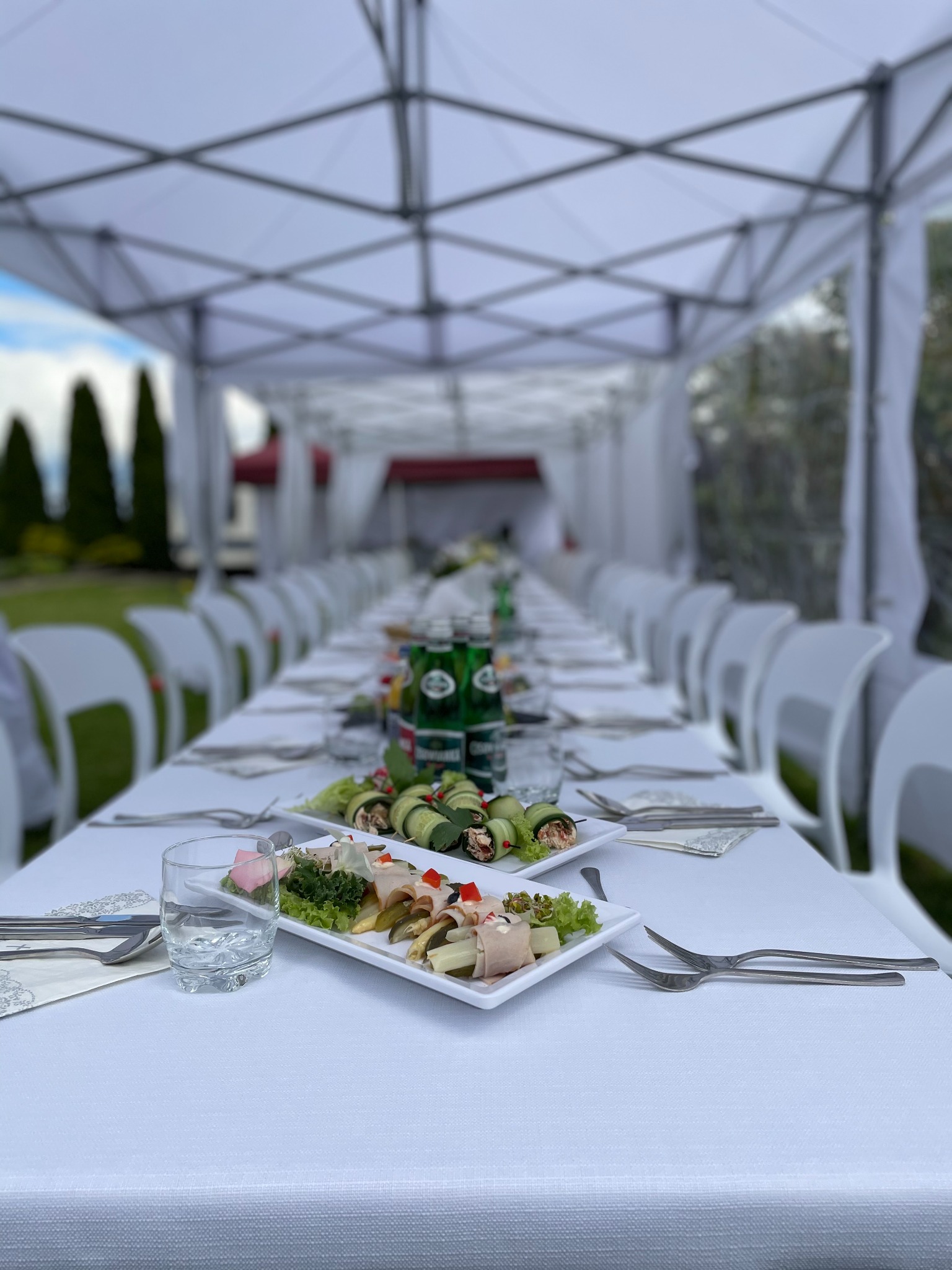 Długi stół cateringowy pod białym namiotem, nakryty białym obrusem, z talerzami przystawek, napojami i sztućcami, przygotowany na imprezę plenerową.
