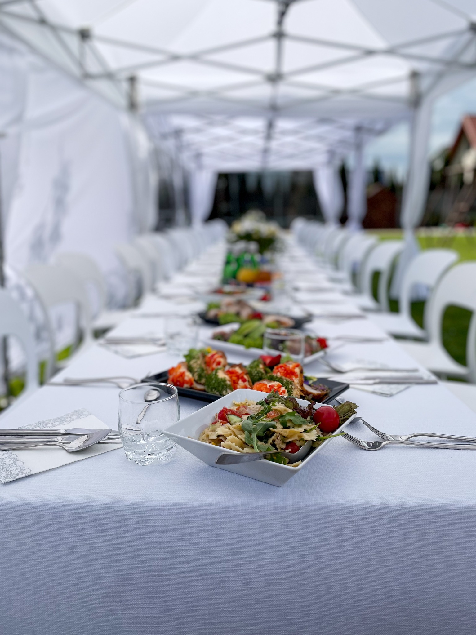 Długi stół przykryty białym obrusem, nakryty zastawą i sałatkami, ustawiony pod białym namiotem.