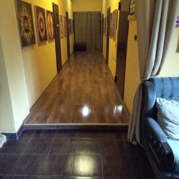 Maciej ALFUTH - Długi korytarz z żółtymi ścianami, na których wiszą obrazy w ciemnych ramach. Podłoga wyłożona brązowymi płytkami przechodzi w drewniany parkiet. Po prawej stronie widoczny fragment sofy z zasłoną.