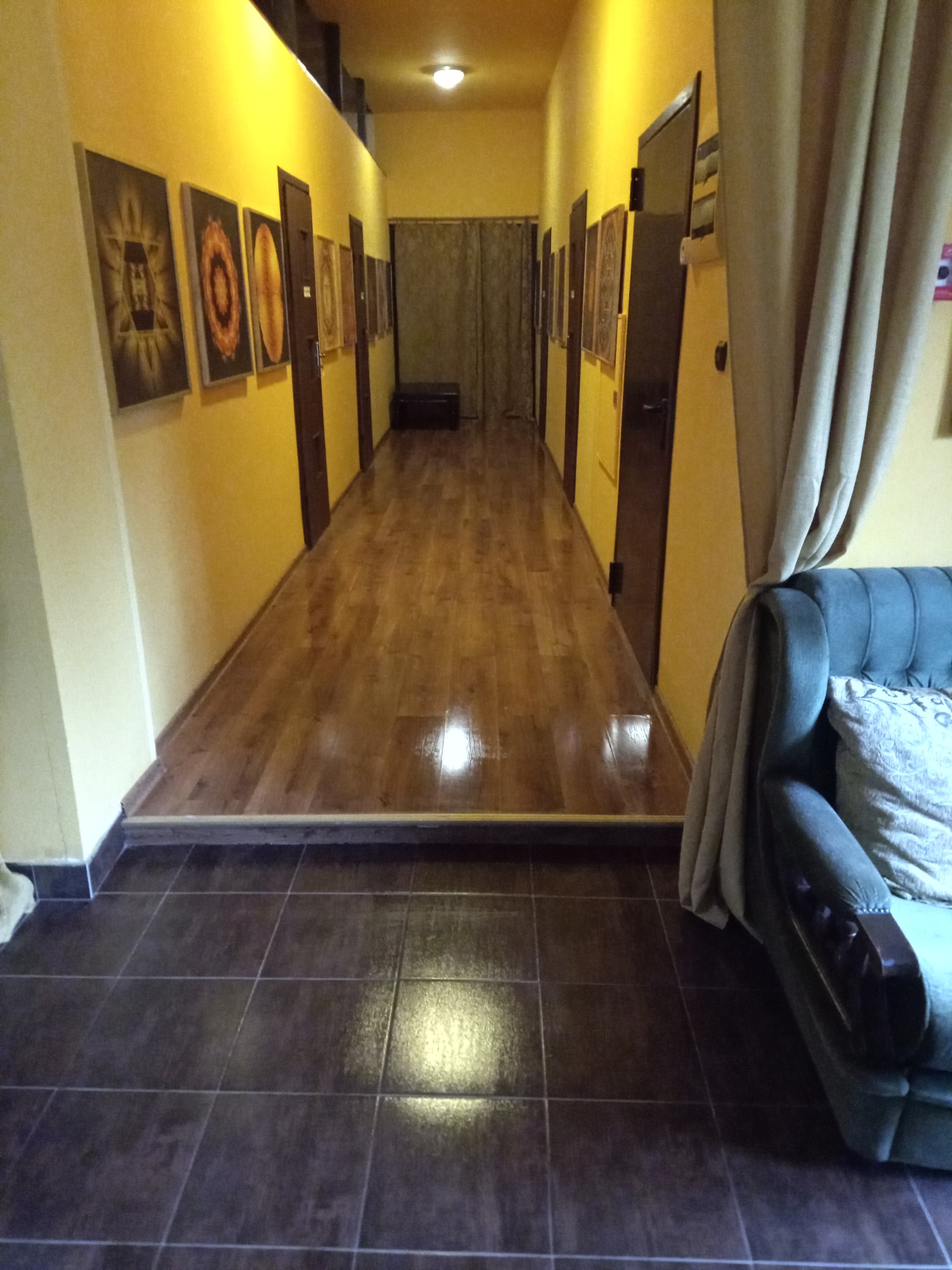 Długi korytarz z żółtymi ścianami, na których wiszą obrazy w ciemnych ramach. Podłoga wyłożona brązowymi płytkami przechodzi w drewniany parkiet. Po prawej stronie widoczny fragment sofy z zasłoną.