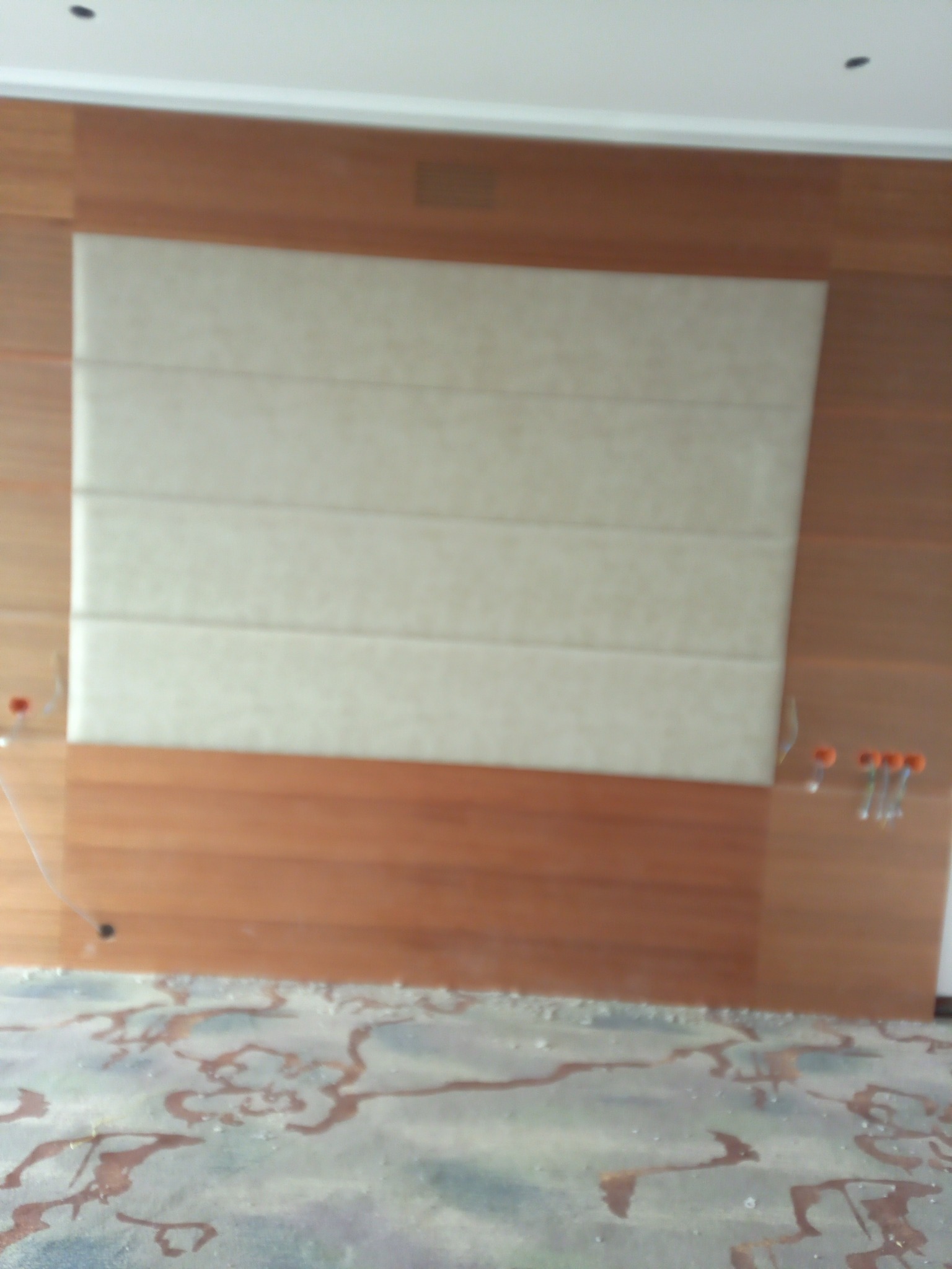 Ściana z drewnianą boazerią i tapicerowanym panelem w kolorze kremowym, widoczne kable elektryczne wystające ze ściany oraz zanieczyszczona podłoga.