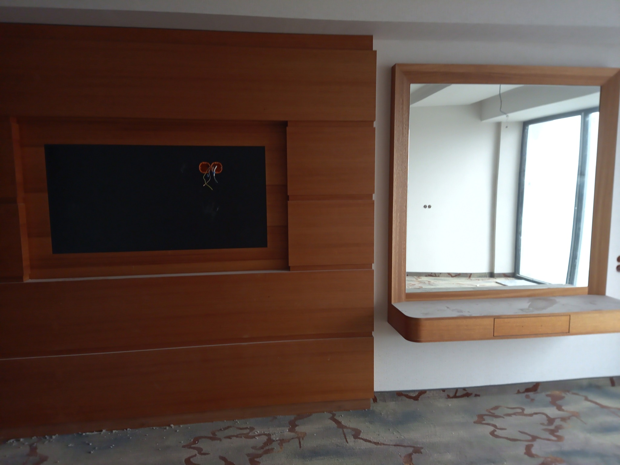 Drewniana zabudowa ściany z czarną wnęką na telewizor i prostokątnym lustrem z półką i szufladą, widoczne przewody elektryczne, podłoga w trakcie prac wykończeniowych.