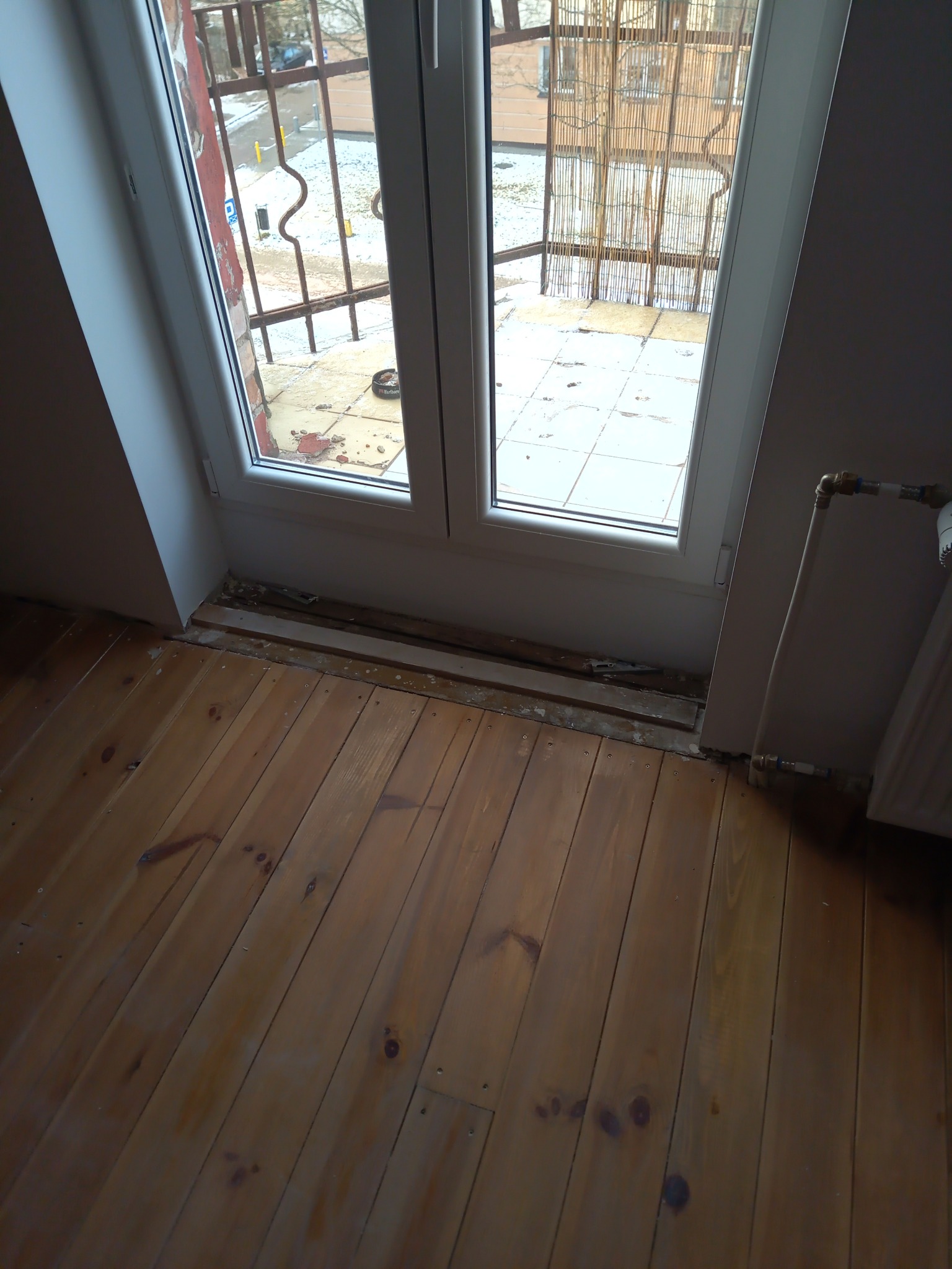 Fragment pokoju na poddaszu z drewnianą podłogą, białym oknem balkonowym i grzejnikiem; widoczne ślady po demontażu listew przypodłogowych.