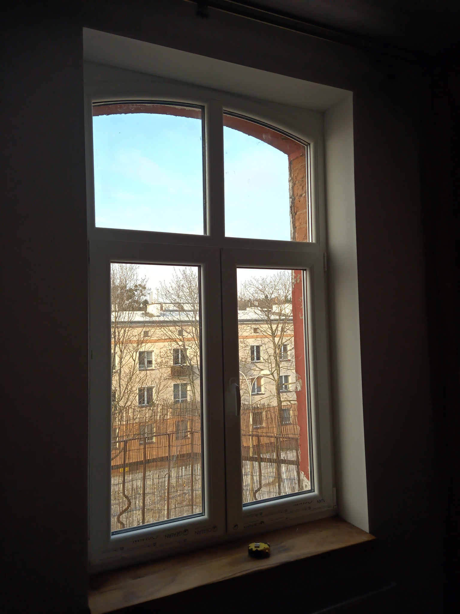 Białe okno PCV z widokiem na budynek mieszkalny i niebieskie niebo, na drewnianym parapecie leży żółta miarka zwijana, ściana w ciemnym odcieniu.