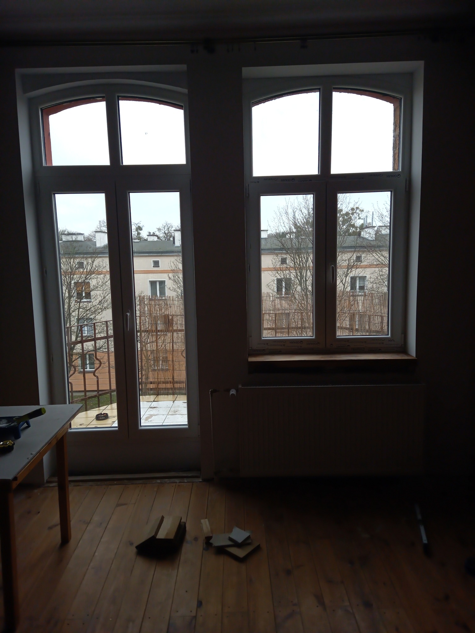 Pokój z drewnianą podłogą w trakcie prac, widoczne dwa okna z widokiem na budynek, stolik z narzędziami i fragmenty materiałów budowlanych na podłodze.