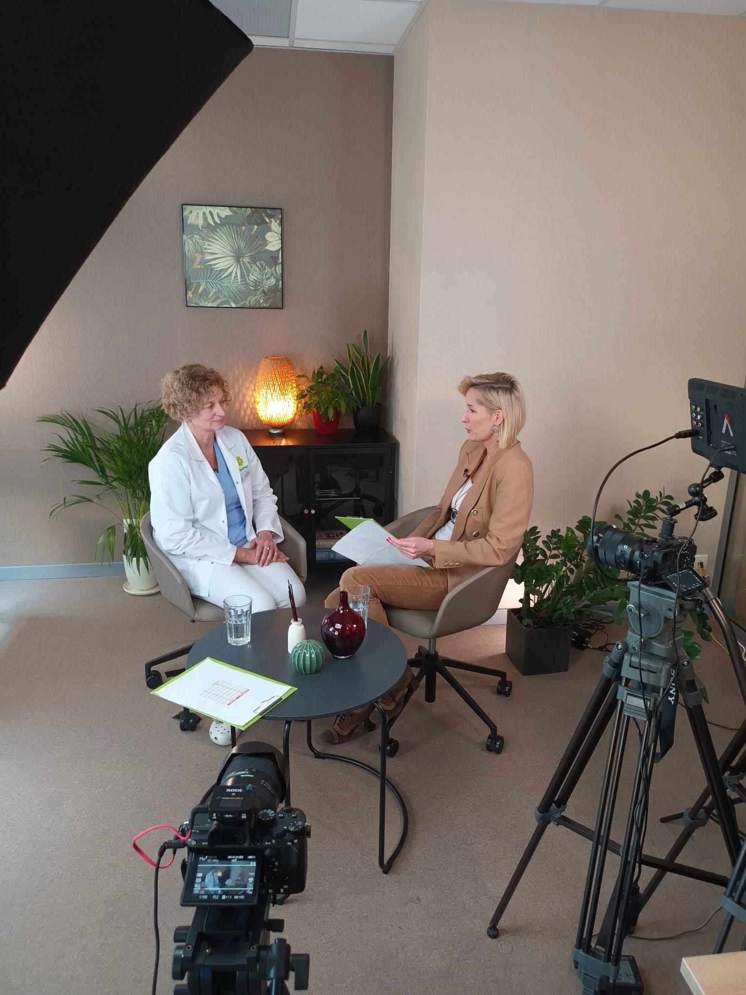Nagranie wywiadu z lekarzem w studio: kobieta w kitlu siedzi naprzeciwko dziennikarki, kamery i oświetlenie ustawione do filmowania rozmowy. W tle rośliny doniczkowe.