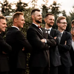 Grupowe portretowe ujęcie uśmiechniętych mężczyzn w garniturach, z których jeden ma czerwoną muchę, stojących ramię w ramię na tle zieleni.