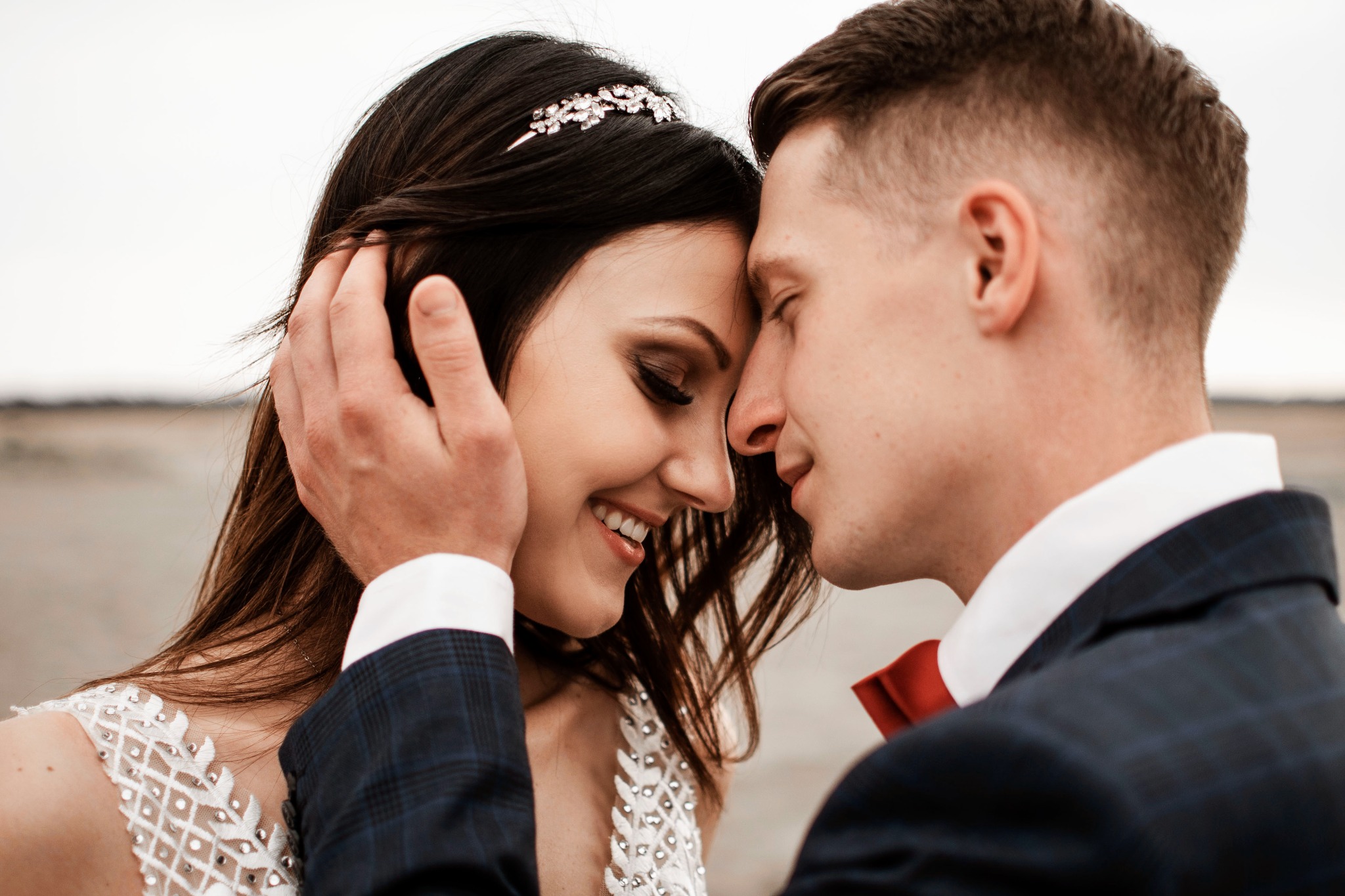 Młoda para, zbliżone ujęcie twarzy, panna młoda z ozdobą we włosach, pan młody w granatowym garniturze z czerwoną muchą, delikatny dotyk dłoni.