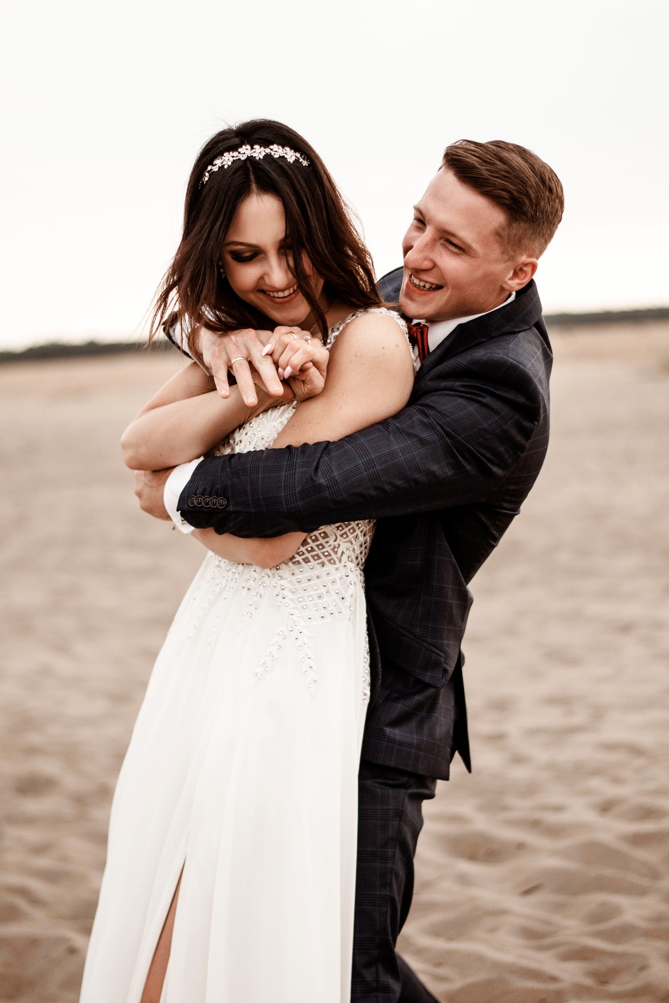 Uśmiechnięta panna młoda w białej sukni z tiulowym dołem i ozdobnym paskiem na głowie, obejmowana przez uśmiechniętego pana młodego w granatowym garniturze w kratę z czerwoną muchą na tle...