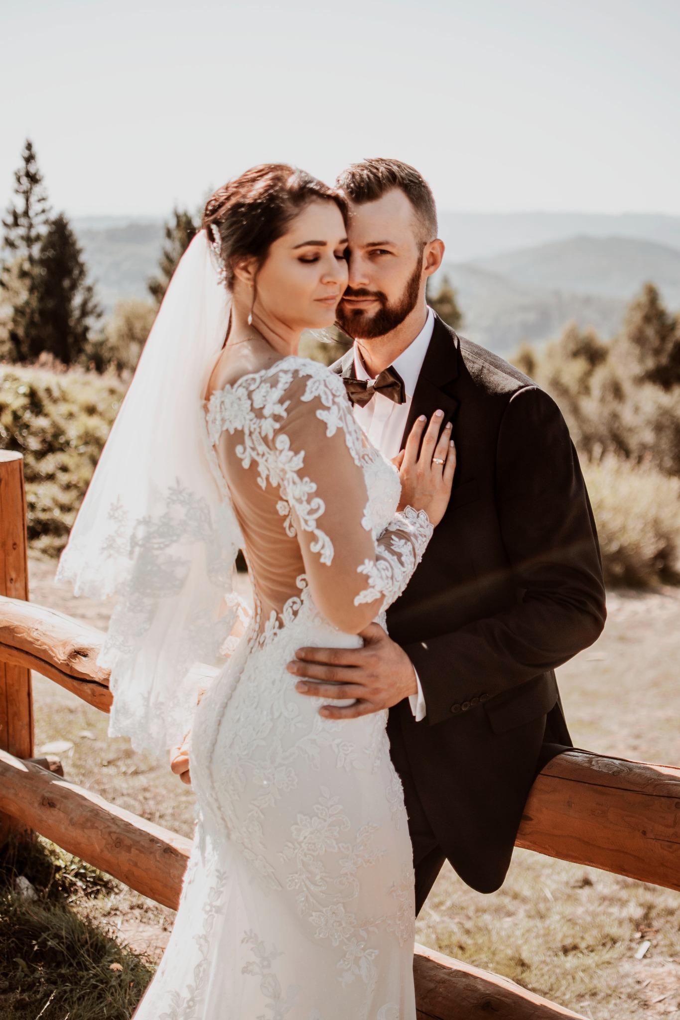 Para młoda przytulona przy drewnianej barierce z widokiem na góry w tle, panna młoda w koronkowej sukni z odkrytymi plecami i welonem, pan młody w czarnym garniturze z muchą, delikatne oświetlenie.