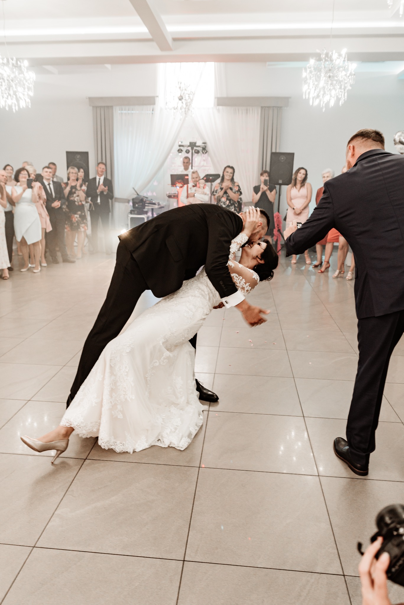 Panna młoda w koronkowej sukni ślubnej, pochylona w pocałunku przez pana młodego podczas pierwszego tańca, w tle zespół muzyczny i goście.