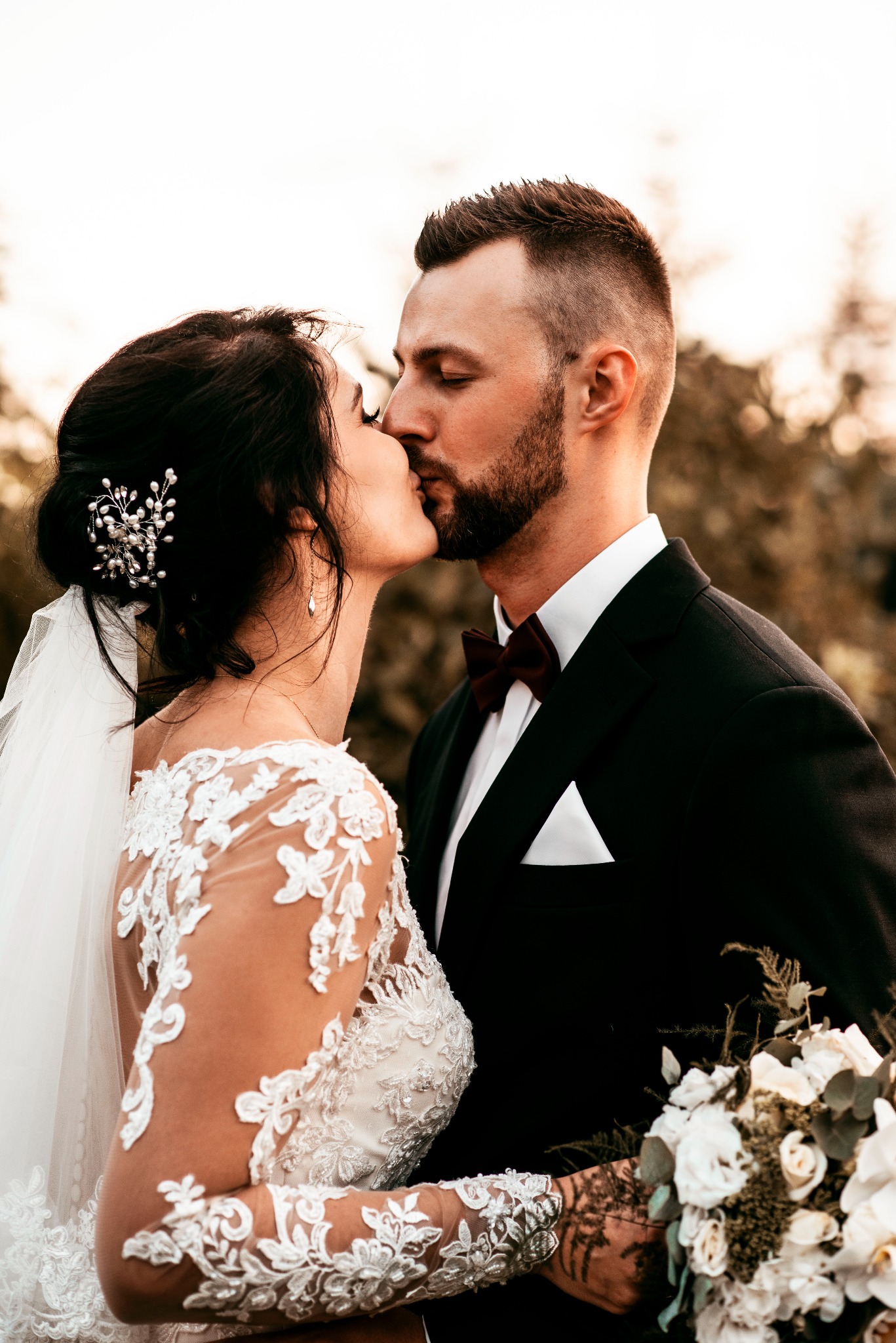 Pocałunek młodej pary; panna młoda w koronkowej sukni z welonem i ozdobą we włosach, pan młody w czarnym smokingu z bordową muchą, trzyma bukiet białych kwiatów.