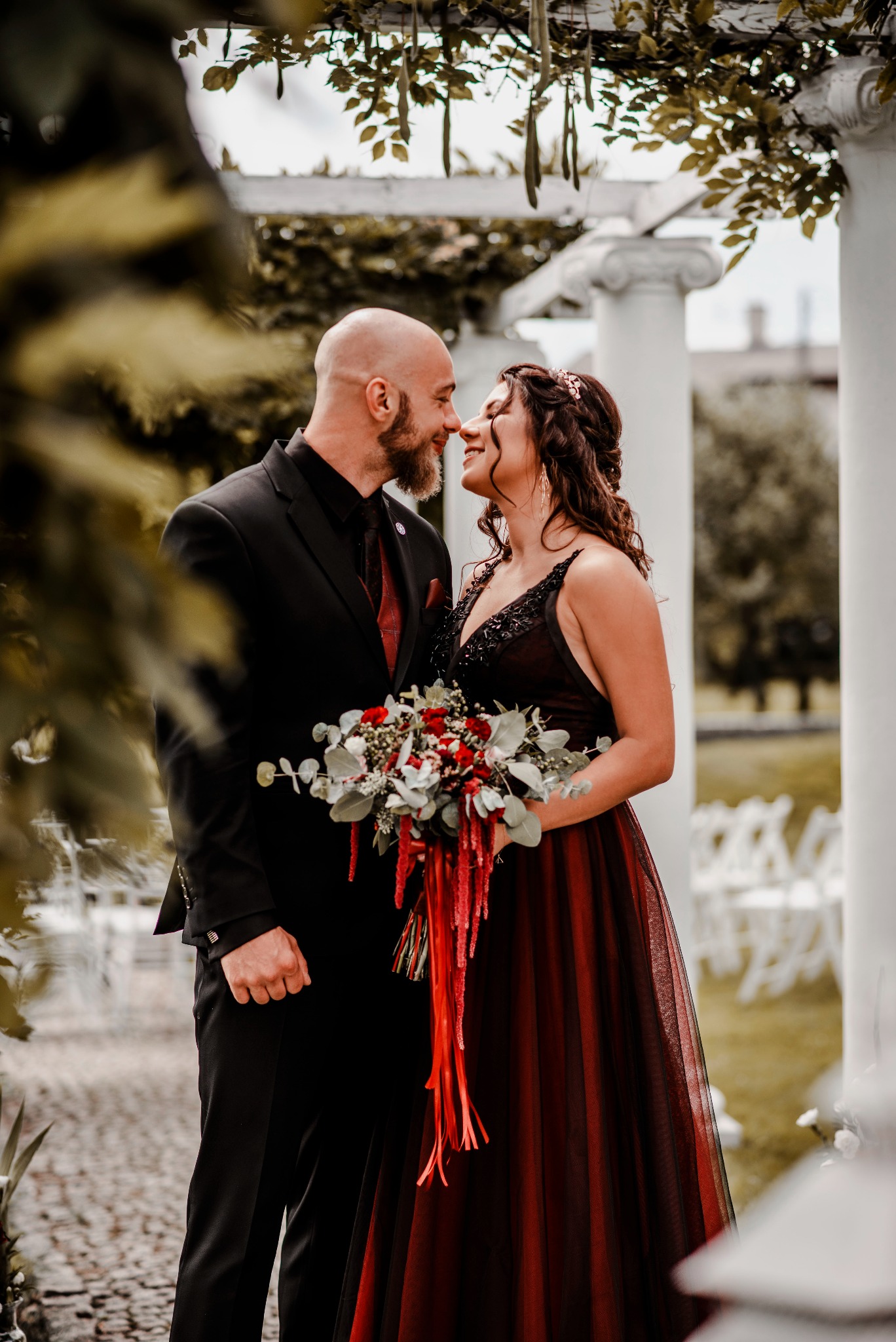 Para młoda, on w czarnym garniturze z bordowym krawatem, ona w bordowej sukni z bukietem czerwonych kwiatów, stoją naprzeciwko siebie pod pergolą z białymi kolumnami, dotykając się nosami.