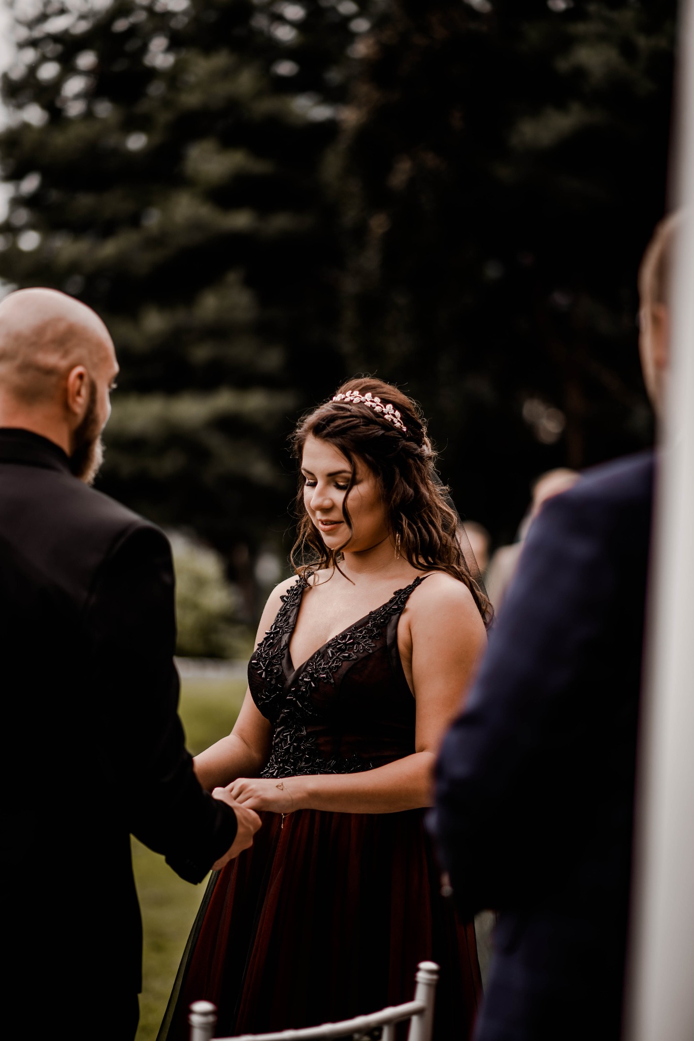 Panna młoda w bordowej sukni, trzymająca dłoń pana młodego podczas ceremonii, z tiarą z motywem liściastym na głowie, na tle zieleni.