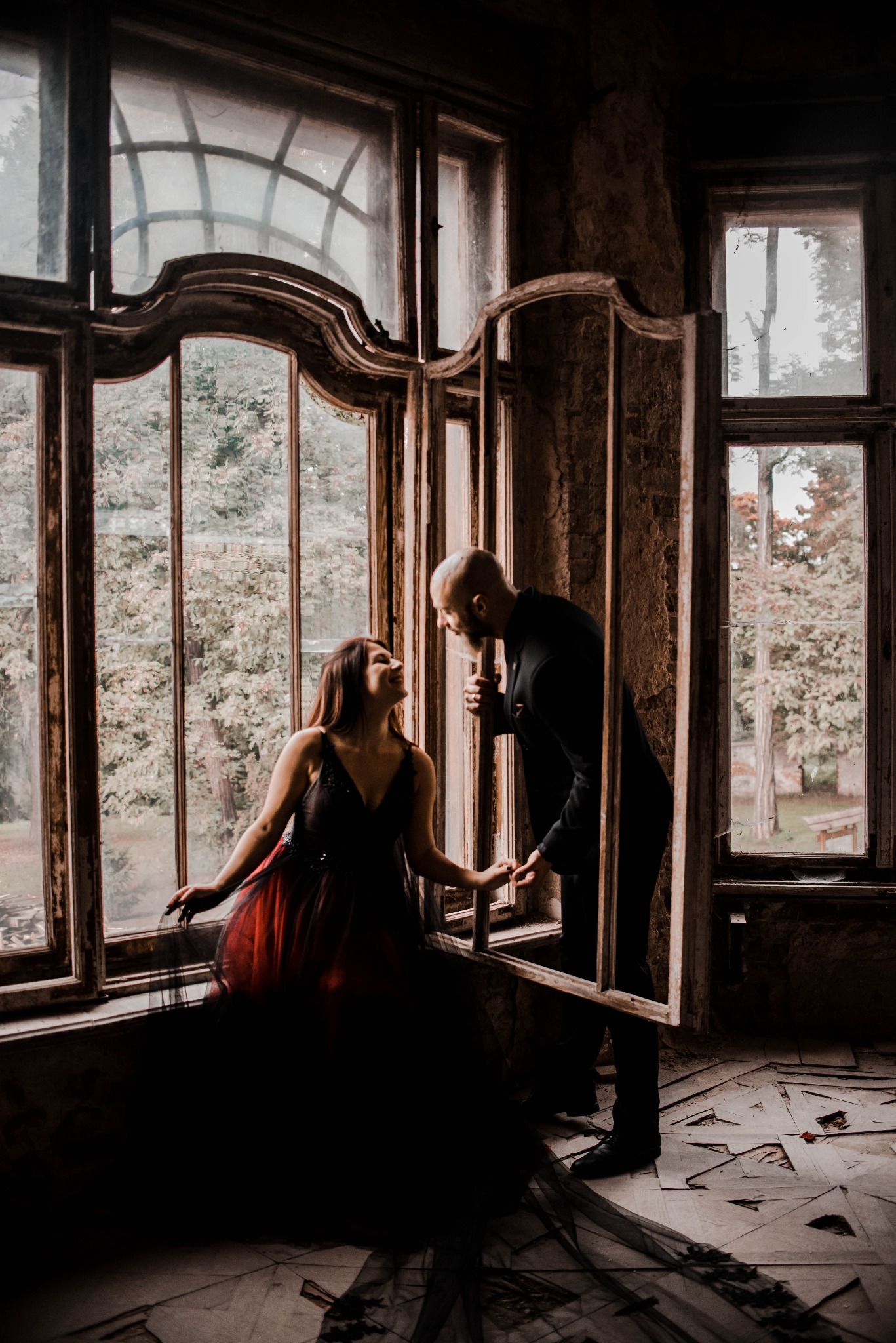 Para w opuszczonym wnętrzu, kobieta w sukni z czerwonym dołem siedzi na parapecie, mężczyzna w czarnym garniturze stoi obok, trzymając ramę okienną, oboje patrzą na siebie.