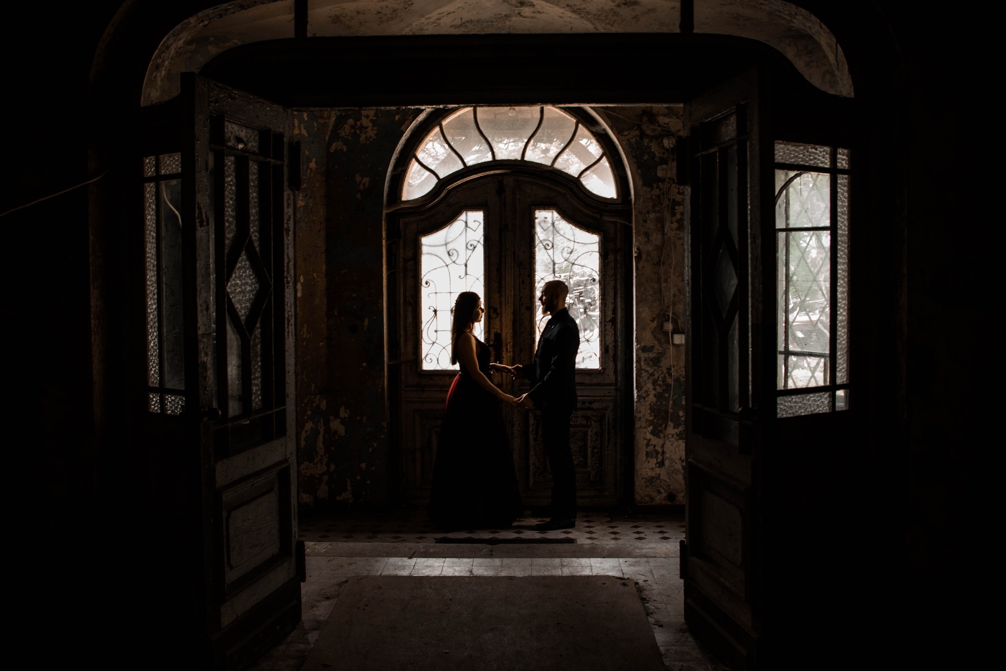 Sylwetki pary trzymającej się za ręce na tle starych, przeszklonych drzwi w opuszczonym budynku z łuszczącą się farbą na ścianach.