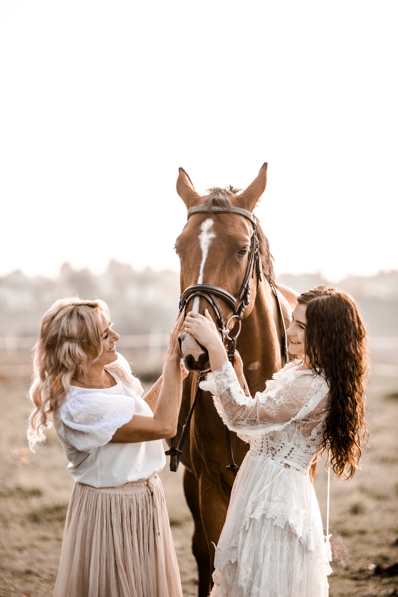 Dwie kobiety w jasnych sukienkach głaszczą brązowego konia z białym znaczeniem na czole, na tle rozmytego krajobrazu w ciepłym świetle.