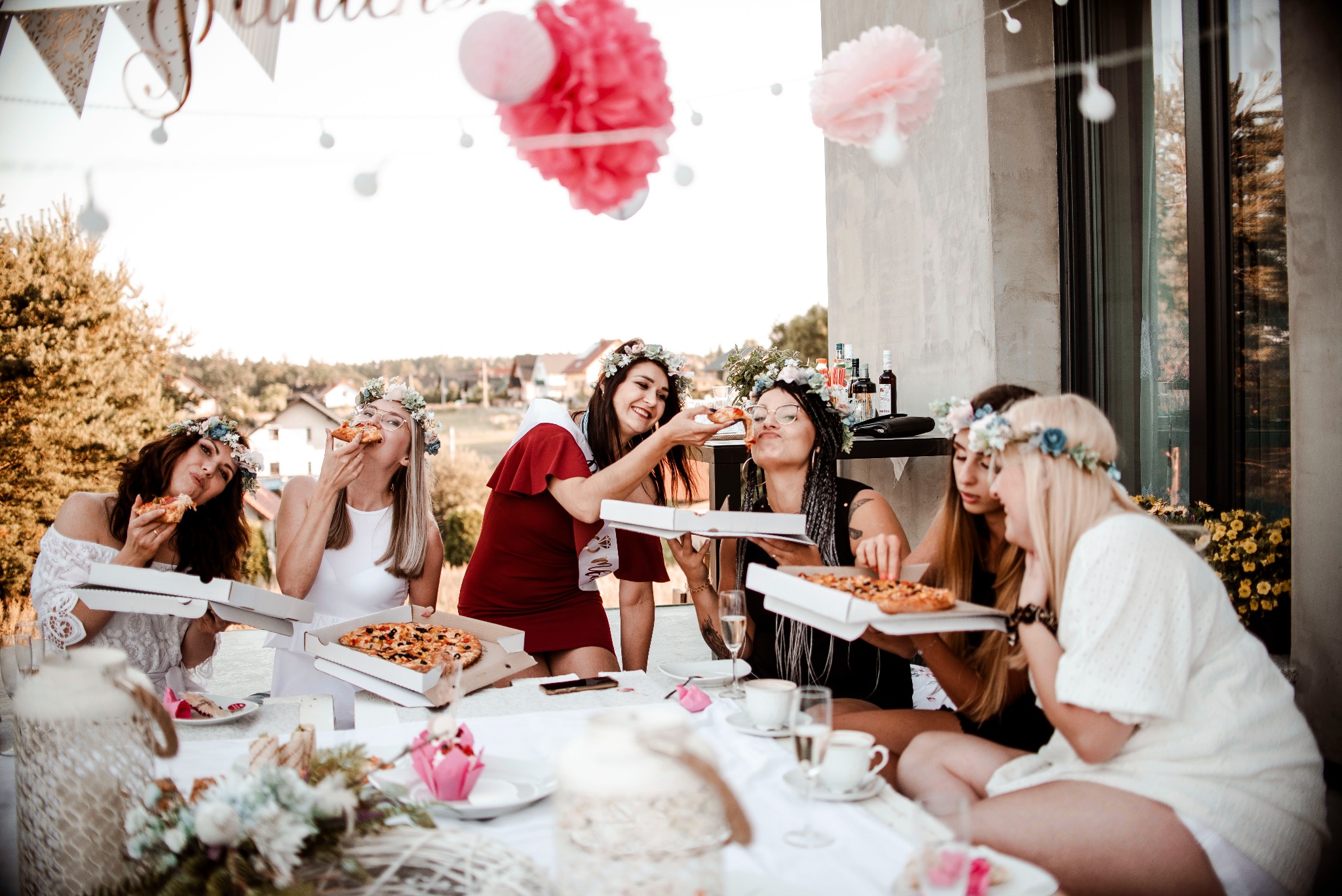 Grupa kobiet w wiankach na głowach siedzi przy stole na zewnątrz, jedząc pizzę i świętując, prawdopodobnie wieczór panieński, dekoracje w tle.