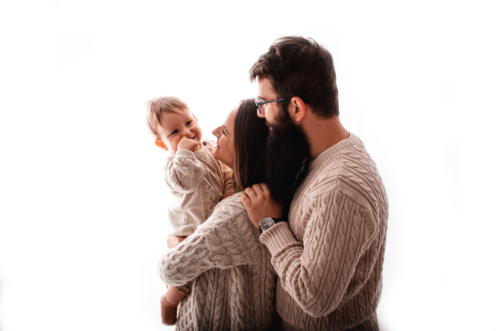 Rodzina w beżowych swetrach: uśmiechnięta matka trzyma na rękach roześmianego synka, obok stoi ojciec w okularach i z brodą, na białym tle.