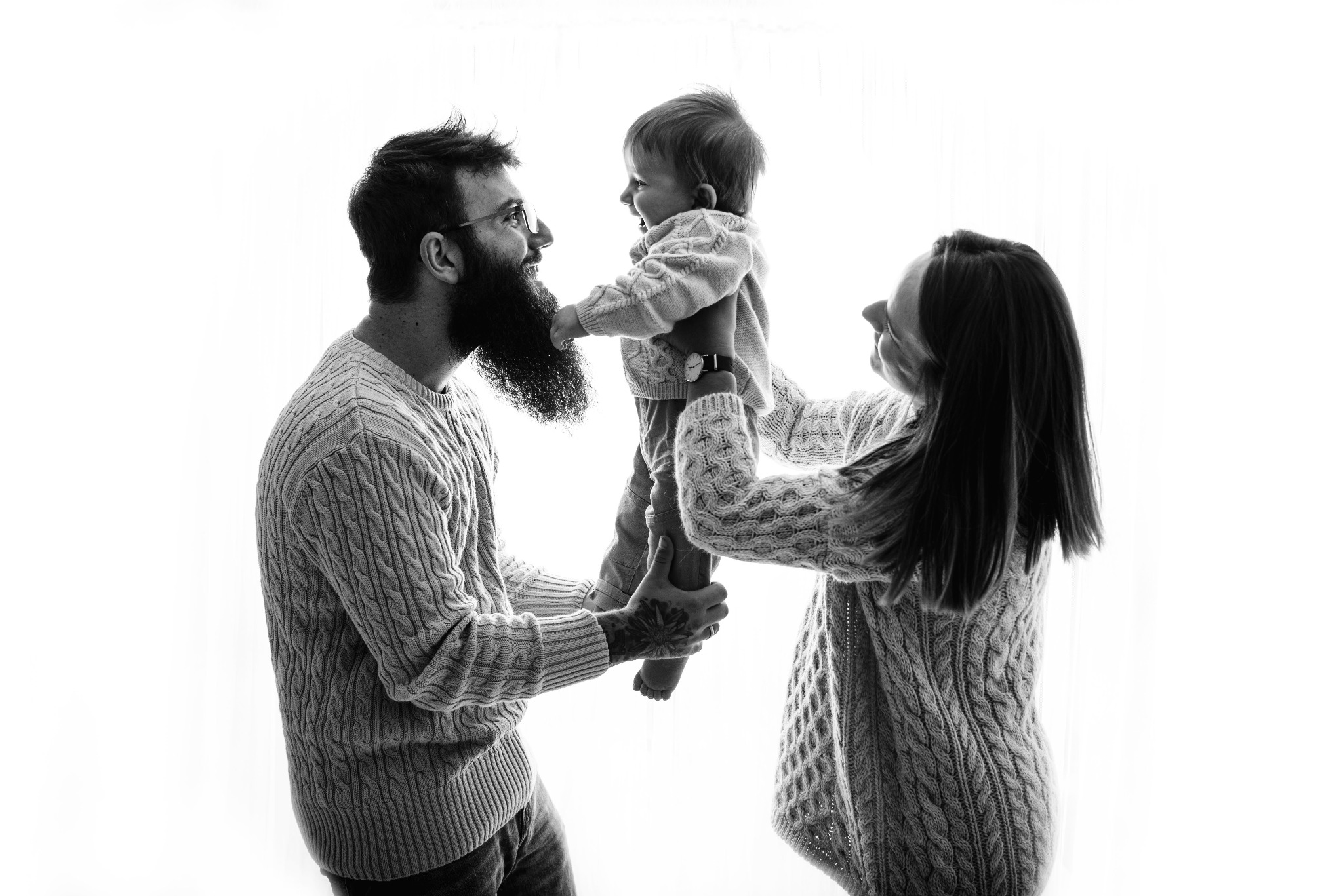 Czarno-białe zdjęcie uśmiechniętej rodziny: ojciec z brodą i tatuażem na dłoni trzyma w górze małe dziecko, matka stoi obok, wszyscy ubrani w swetry z warkoczowym splotem, na jasnym tle.