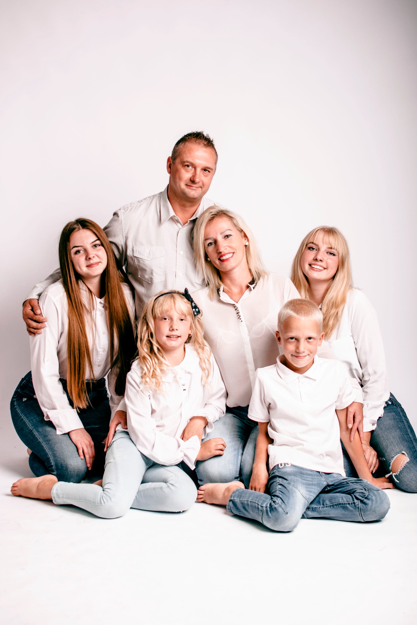 Rodzinna sesja portretowa: rodzice z czwórką dzieci w jasnych ubraniach na białym tle, uśmiechnięci, siedzą na podłodze.