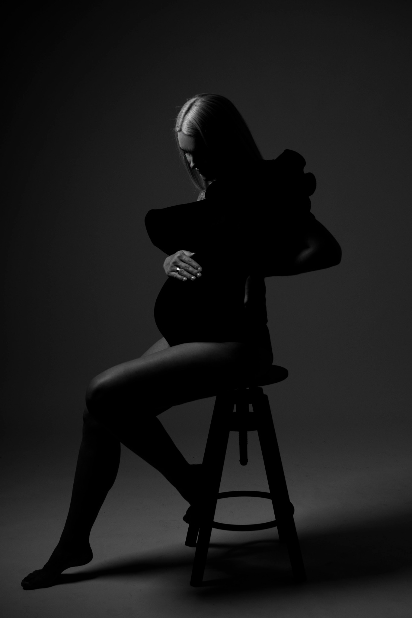 Czarno-białe zdjęcie sylwetki kobiety w ciąży, siedzącej na stołku, z rękami na brzuchu, w ciemnym otoczeniu studyjnym.