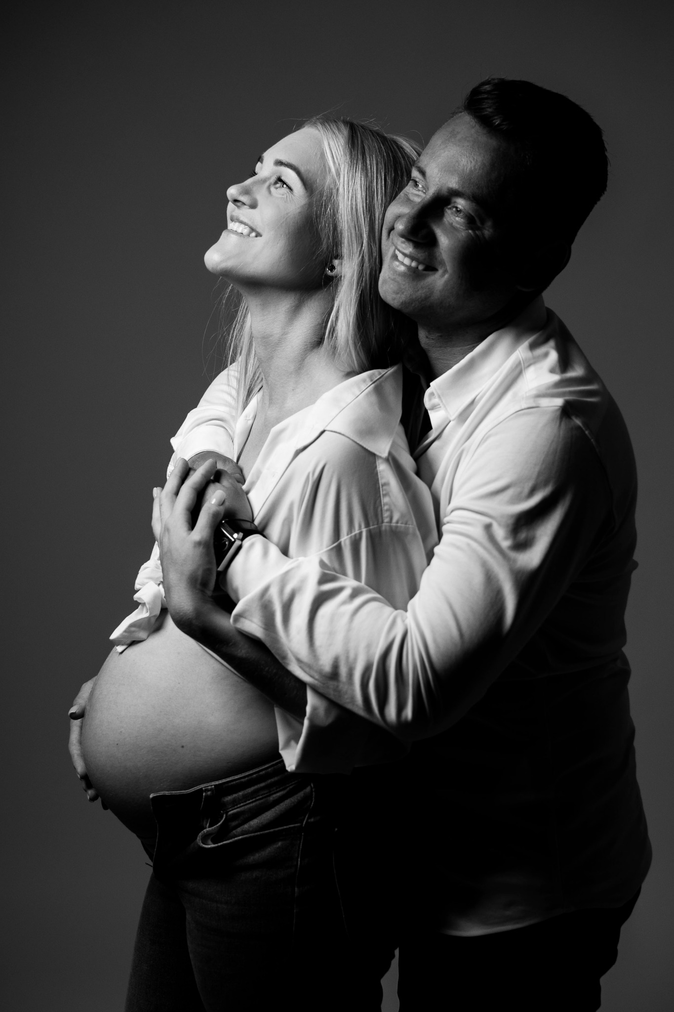 Czarno-białe zdjęcie obejmującej się pary: uśmiechnięta, ciężarna blondynka w rozpiętej koszuli i dżinsach, oraz mężczyzna w koszuli, oboje patrzą w górę.