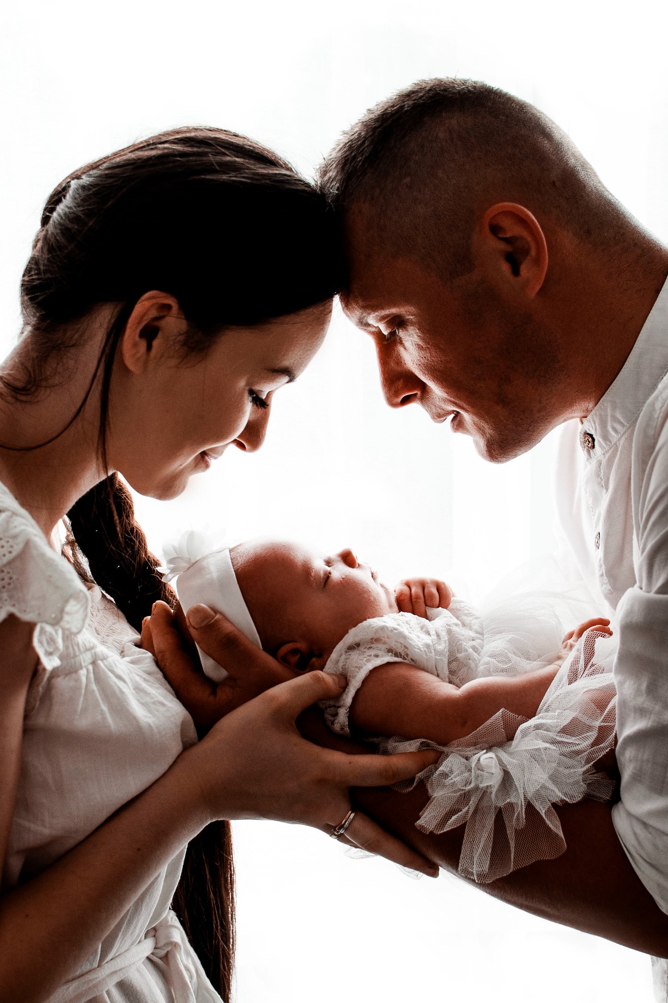 Rodzice spoglądają z czułością na śpiące nowonarodzone dziecko ubrane w białą koronkową sukienkę i opaskę, trzymane w ramionach, z widocznym pierścionkiem na palcu matki.