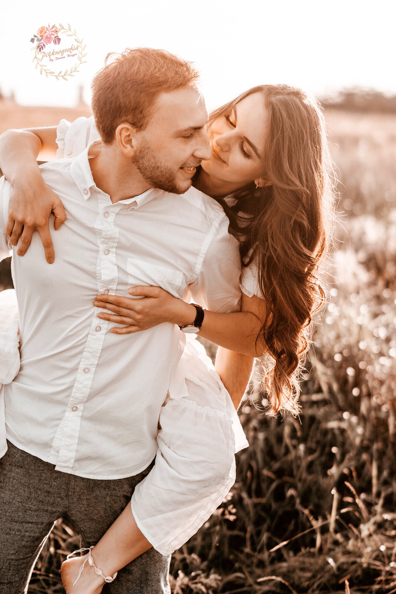 Uśmiechnięta kobieta obejmuje mężczyznę w białej koszuli na tle pola w ciepłym, zachodzącym słońcu. Kobieta ma długie, brązowe włosy i delikatną bransoletkę na kostce.