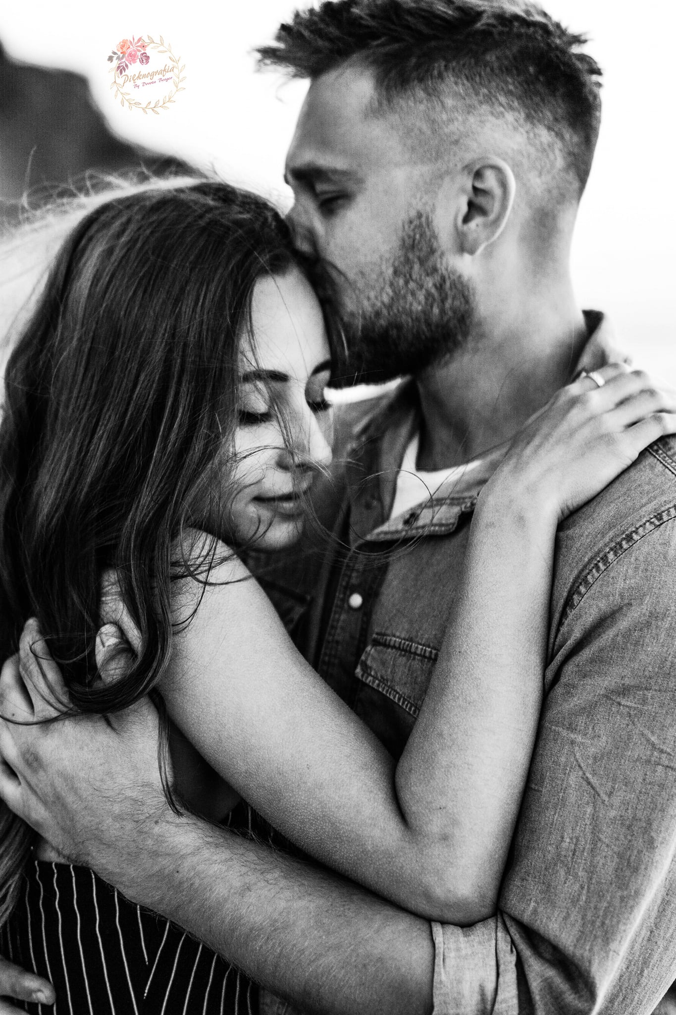 Czarno-białe zdjęcie obejmującej się pary: mężczyzna całuje kobietę w czoło, ona ma zamknięte oczy, wiatr rozwiewa jej włosy.