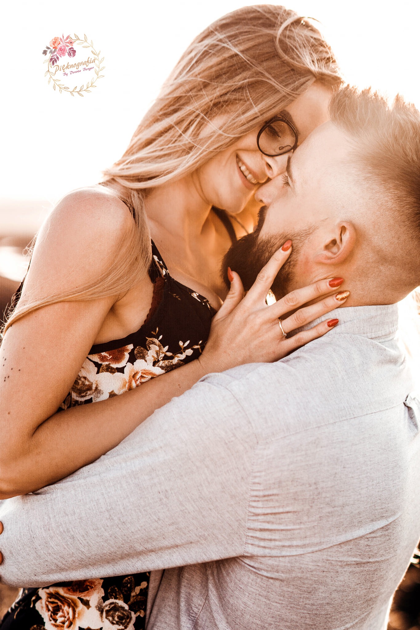 Uśmiechnięta kobieta w okularach i sukience w kwiaty obejmuje brodatego mężczyznę, dłoń z pierścionkiem na jego twarzy, ciepłe światło.