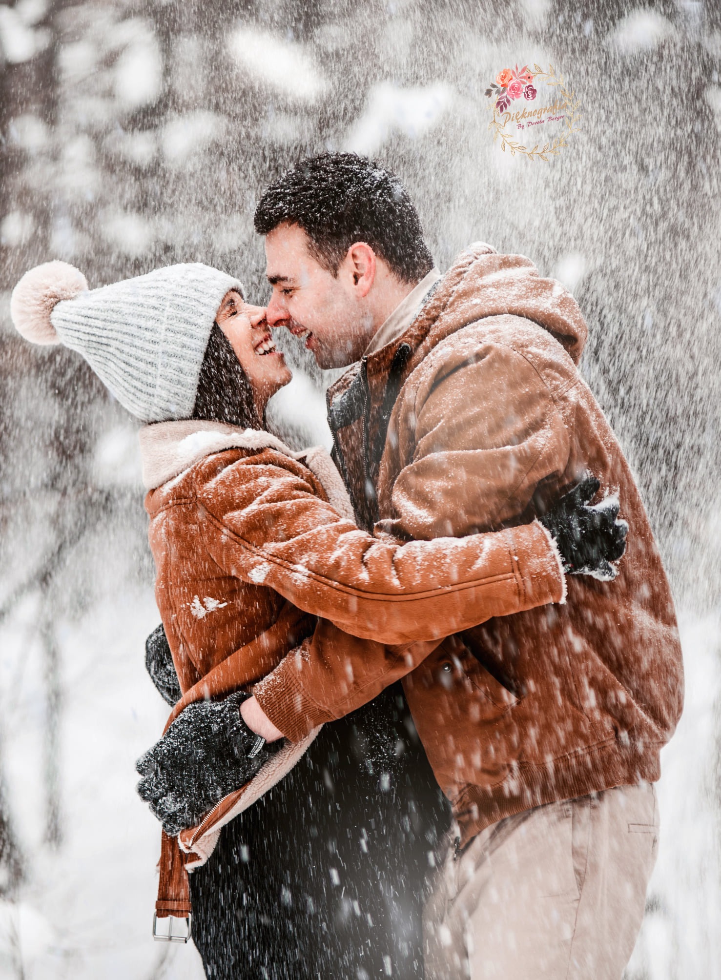 Uśmiechnięta para w brązowych kurtkach, obejmująca się w śnieżny dzień, z widocznym logo fotografa w prawym górnym rogu.