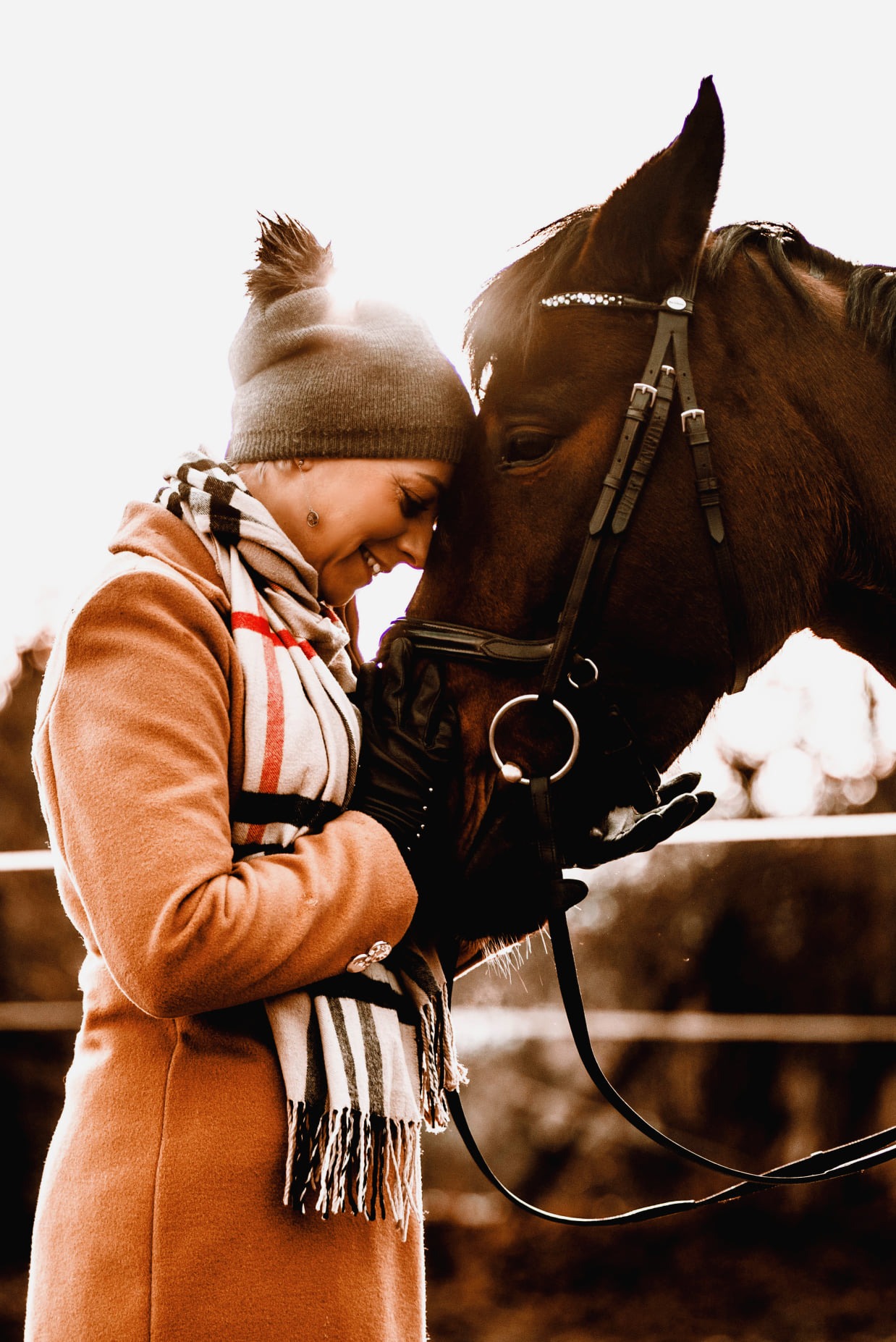 Uśmiechnięta kobieta w płaszczu i czapce przytula głowę brązowego konia z ozdobnym ogłowiem, delikatne, rozproszone światło.
