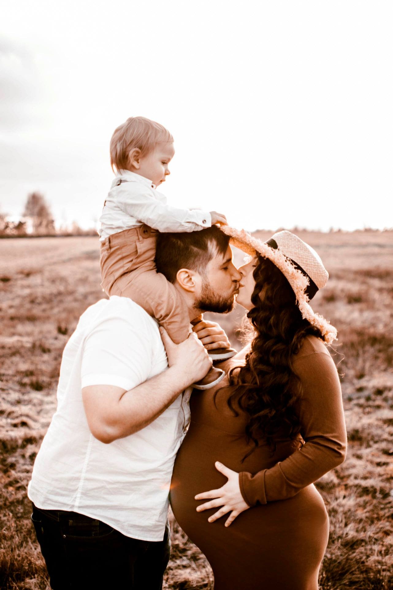 Przyszła mama w brązowej sukience i kapeluszu całuje się z partnerem, na którego ramionach siedzi ich mały syn na tle łąki o zachodzie słońca.